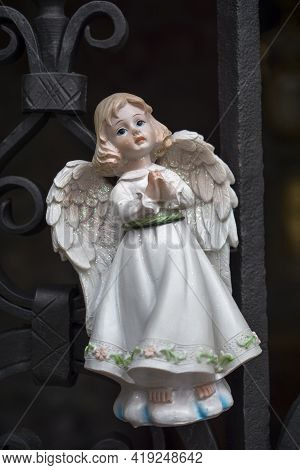 Figurine Of An Angel In A White Dress. Decorative Figure On A Metal Railings Background. Statuette. 