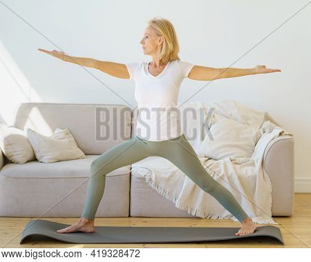 Calm And Concentrated Senior Woman In Home Wear Practicing Warrior Yoga Pose While Exercising In Mor