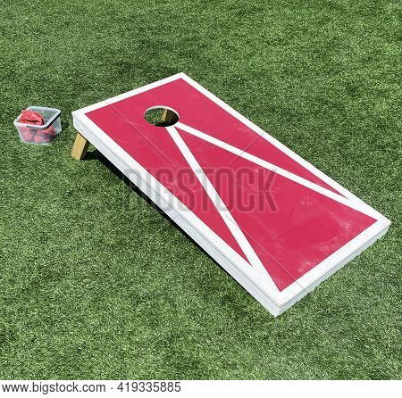Red And White Bean Bag Toss Cornhole Game On A Green Turf Field.