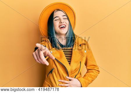 Young modern girl wearing yellow hat and leather jacket laughing at you, pointing finger to the camera with hand over body, shame expression 