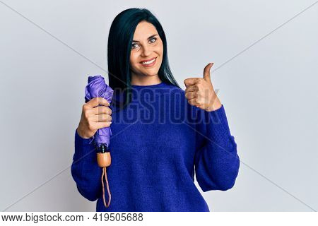 Young caucasian woman holding purple closed umbrella smiling happy and positive, thumb up doing excellent and approval sign 