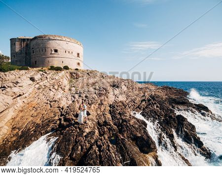 Man And Woman In Love Stand Embracing And Holding Hands On The Rocky Seashore Near Arza Fortress
