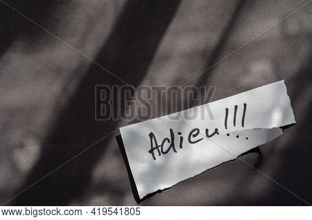 Handwritten Word Adieu On A White Piece Of Paper. A Scrap Of Paper With A Jagged Edge On A Gray Back