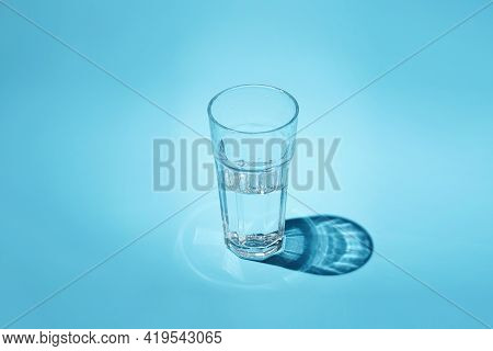 A Glass Of Water - Half Full Or Half Empty - In Hard Light With Nice Shadow On Blue Surface