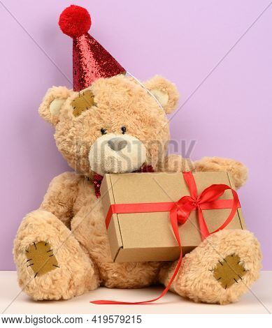 Cute Brown Teddy Bear In A Red Cap Sits And Holds A Brown Box With A Gift, Festive Background