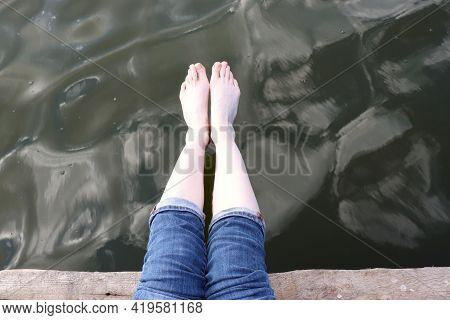 Woman Barefoot Legs In Blue Jeans On Wooden Pier On River Water Background. Summer Vacation Concept.