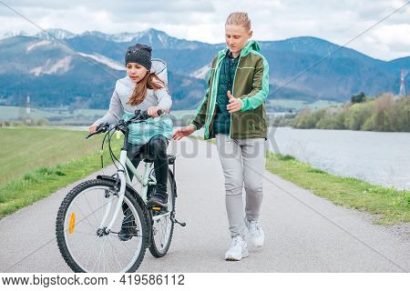 Laughing Kids Have Fun Using A Skateboard. Brother Pushing His Sister Sitting On A Skateboard And Ma