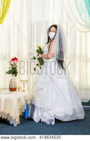 A Jewish Bride Sits Waist-deep In A Synagogue At A Table With Flowers Before A Chuppa Ceremony Durin