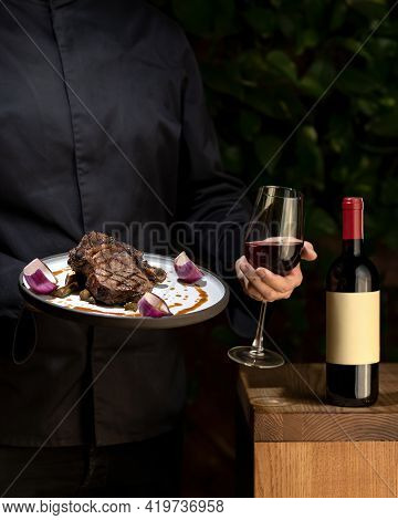Glass Of Red Wine And Plate With Cooked Meat In Hands Of Unrecognizable Person On Black Background. 