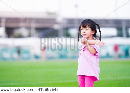 Cute Asian Girl Is Exercising In The Grass. Happy Child Is Using Both Arms And Twists The Body On Th