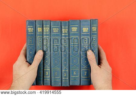 Books Of Classical Literature On A Red Background. Men's Hands Hold The Collected Works Of Jules Ver