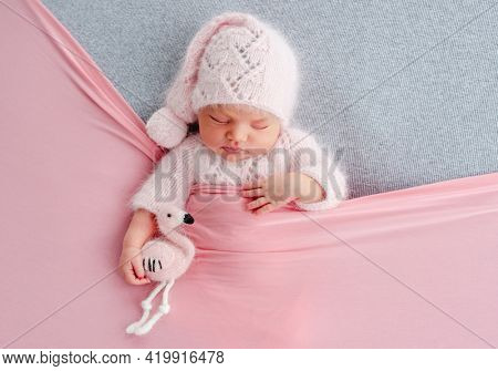 Cute newborn baby girl in knitted hat sleeping on grey stylized background and holding flamingo toy in her hands. Adorable infant child napping under pink blanket during studio photoshoot