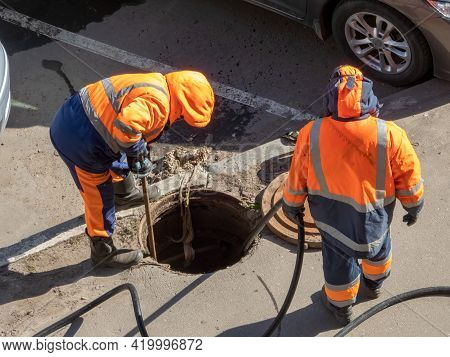 Workers Over The Open Sewer Hatch On A Street. Repair Of Sewage, Underground Utilities, Water Supply