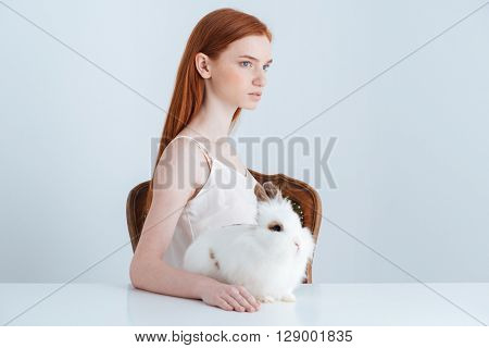 Pensive redhead woman sitting at the table with rabbit and looking away isolated on a white background