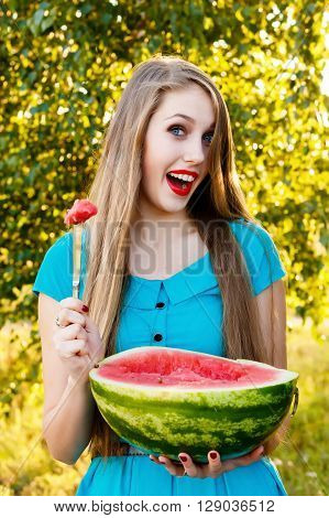 Beautiful blonde girl in a blue dress with long hair eating a watermelon outdoors