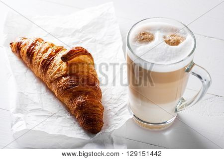 Glass of latte macchiato and French croissant on baking paper. Horizontal scene on white wooden background.
