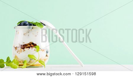 Yogurt oat granola with jam, blueberries and green leaves in glass jar on pastel mint backdrop, copy space
