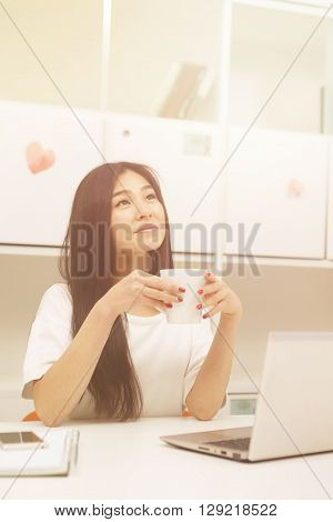 Portrait of Asian student dringking cup of tea or coffee while studying in library with laptop computer. Happy brunette lady looking upwards. Toned image.