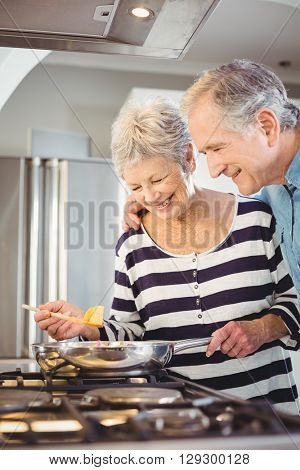 Happy senior man standing with wife cooking food at hob in kitchen