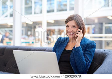 Attractive young businesswoman smiling at the camera, while holding her phone to her ear and sitting on a couch in a modern office space with her laptop computer on her lap