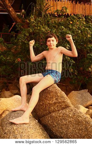 preteen hadsome suntanned boy in swimming slop show biceps on the stone rock beack background close up photo