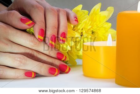 Skin care of a beauty female hands with pink and yellow striped nail art manicure. And yellow chrysanthemums with yellow two candles on the background