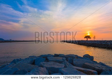 Sunset with colorful sky at a beach in Pattaya Thailand with foreground rocks and view of small trees on rocky peirs against colorful sunset sky