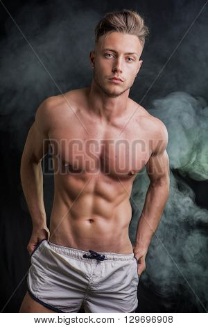 Lean athletic shirtless young man standing on dark background, with smoke around him