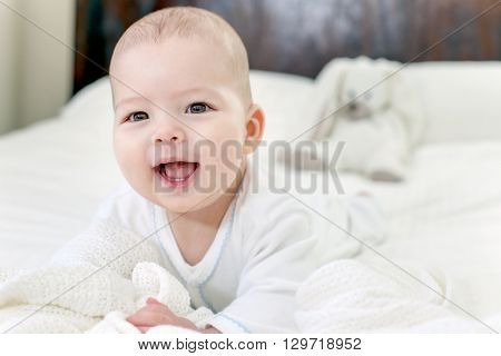 Baby boy wearing white and blue in white sunny bedroom on white bed. Newborn child relaxing on bed after bath or shower. Nursery for children. Textile and bedding for kids. New born kid with toy bunny.
