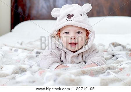 Baby boy wearing a bear outfit in white sunny bedroom on grey blanket. Newborn child relaxing on bed after bath or shower. Nursery for children. Textile and bedding for kids. New born kid with bear. New born kid during tummy time.