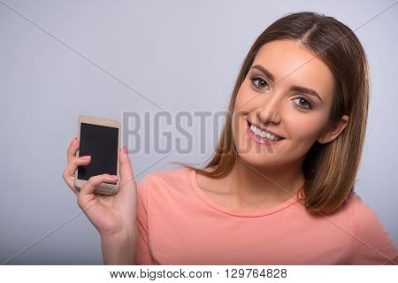 Magic device.  Positive content smiling woman holding cellphone and smiling while standing isolated on grey background