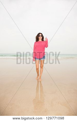 brunette summer vacation woman with sunglasses red sweater blue jeans shorts barefoot standing finger and hand up on sand with ocean behind in beach in Cadiz Andalusia Spain Europe