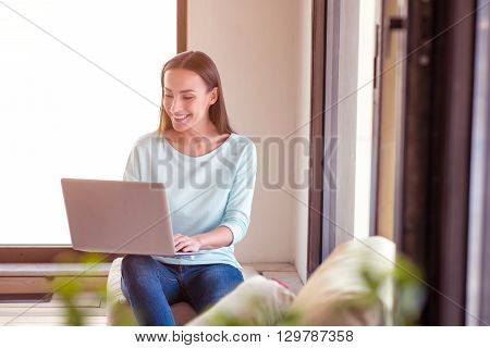 Involved in positivity. Cheerful charming content smiling woman sitting near window and resting while using laptop