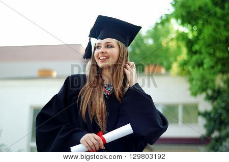 A beautiful graduate girl in the mantle keeps in hands graduate diploma and looks up.