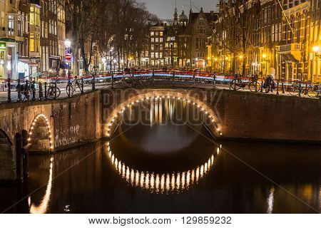A view of the bridges at the Leidsegracht and Keizersgracht canals intersection in Amsterdam at dusk. Bikes and buildings can be seen. The trail of traffic can be seen on a bridge.