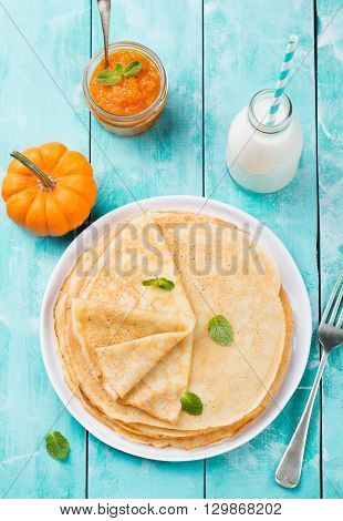 Thin crepes or pancakes with fresh pumpkin confiture, jam and bottle of milk on a rustic wooden background. Top view