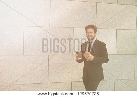 Toned picture of smiling businessman eating junk food and drinkig cupof tea or coffee while walking to office along street.