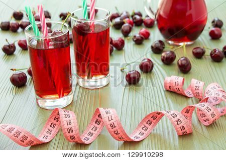 Left 2 glasses of juice with straws, scattered cherries, jar of cherry juice, in front centimeter on light green background. Cherries, cherry juice and centimeter. Horizontal.