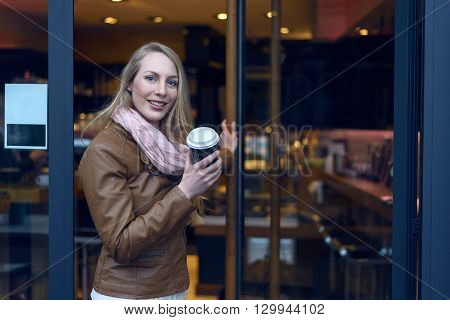 Young blond woman in brown jacket and scarf holding a cup of coffee when leaving the coffeebar