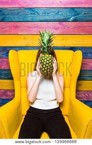 Girl holds pineapple in front of the face. Young girl on a colorful striped background. Sitting fruit fashion girl. Full face. Bright juicy picture