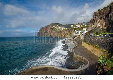 Long Madeiran embankment view and villages on the mountains. Rocky shores of Madeira island south coast. Walking route near Camara de Lobos town.