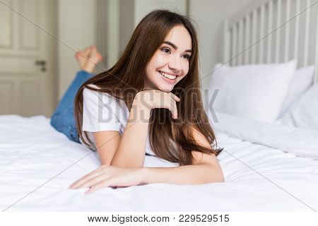 Young Woman Lying At The End Of The Bed Underneath The Quilt And Smiling, With Her Head Resting Upon