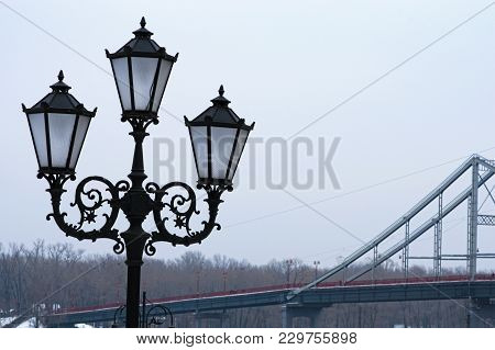 Beautiful Black Street Lantern For Three Lamps, Close Up Photo. Pedestrian Bridge In The Background.