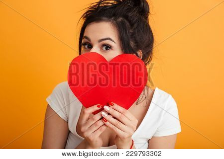 Closeup imege of gorgeous woman 20s in casual clothing covering her face with paper red heart and demonstrating affection over yellow background