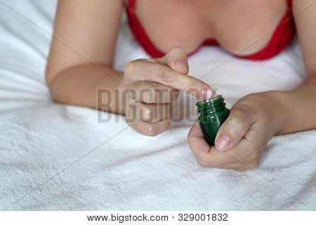 Woman In Red Lingerie Applies Green Herbal Balm From The Bottle To The Skin Lying On The Bed. Concep