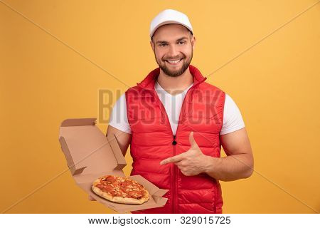 Cheerful Pizza Delivery Man Stands With Carton Boxes, Point To The Pizza, Wears Yellow Hat And White