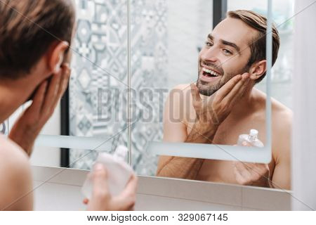 Handsome smiling young shirtless man applying aftershave lotion while standing at the bathroom mirror