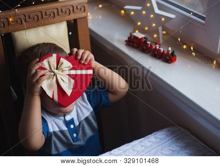 Little Child Boy Hiding Behind Red Heart Shaped Gift Box With Bow Near Window In Daylight. Valentine