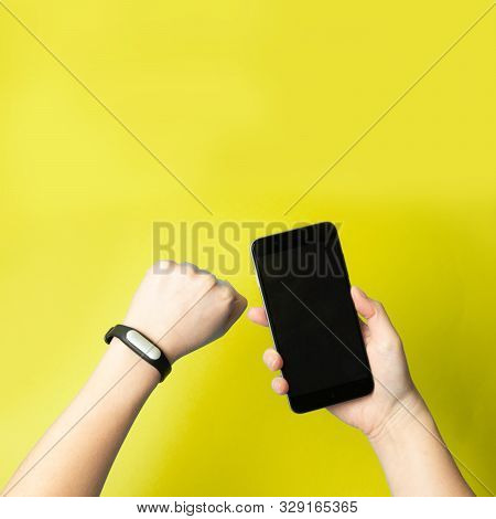Top View Female Hands Hold A Smartphone And A Fitness Bracelet On Hand On A Yellow Background