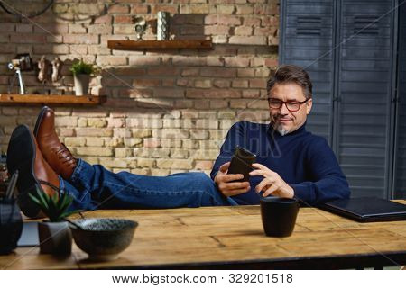 Older white man in glasses sitting at desk in dark room at home with feet on desk, working, using phone.